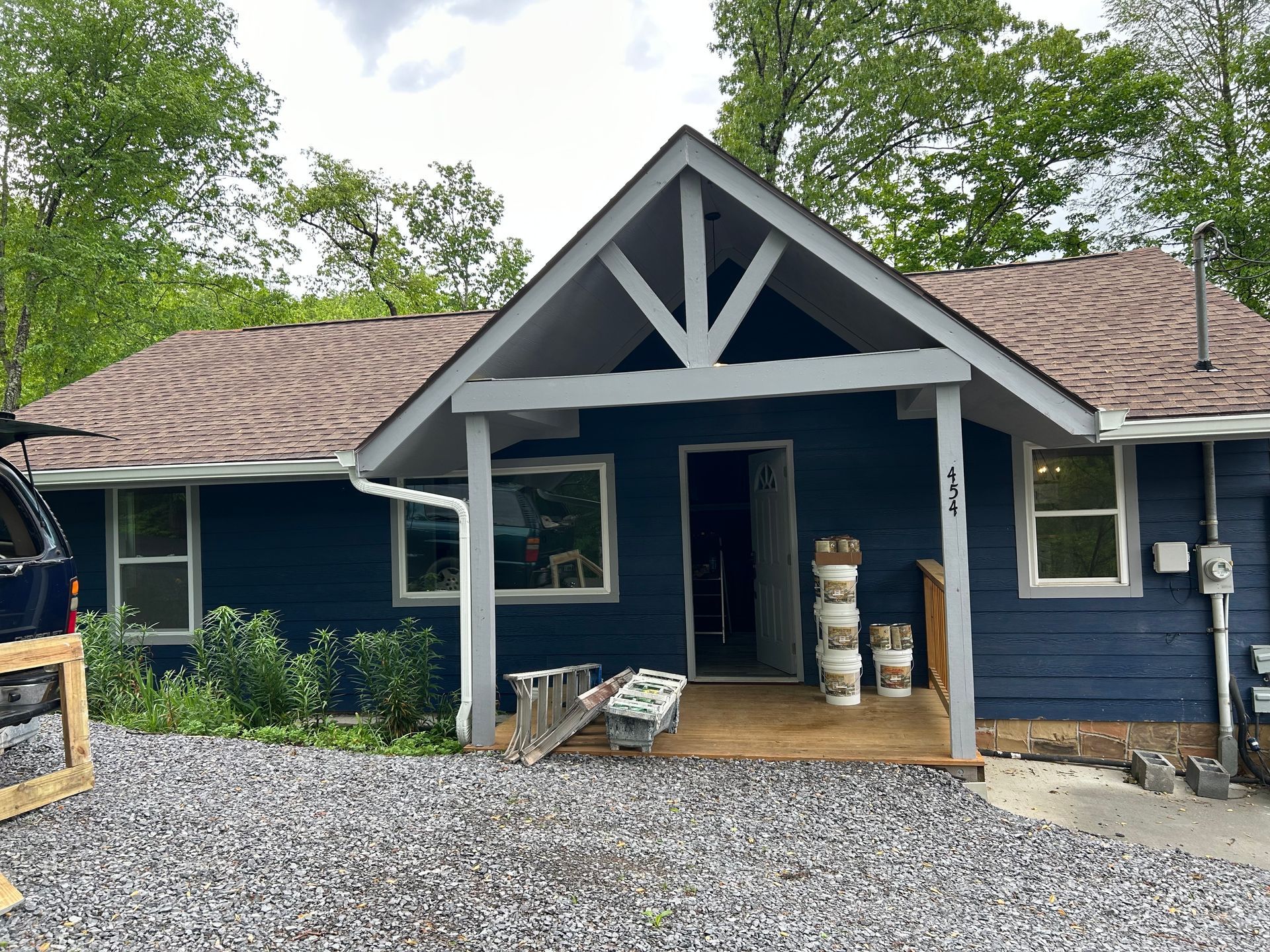 Blue house with white trim, covered porch, and brown roof under a cloudy sky.