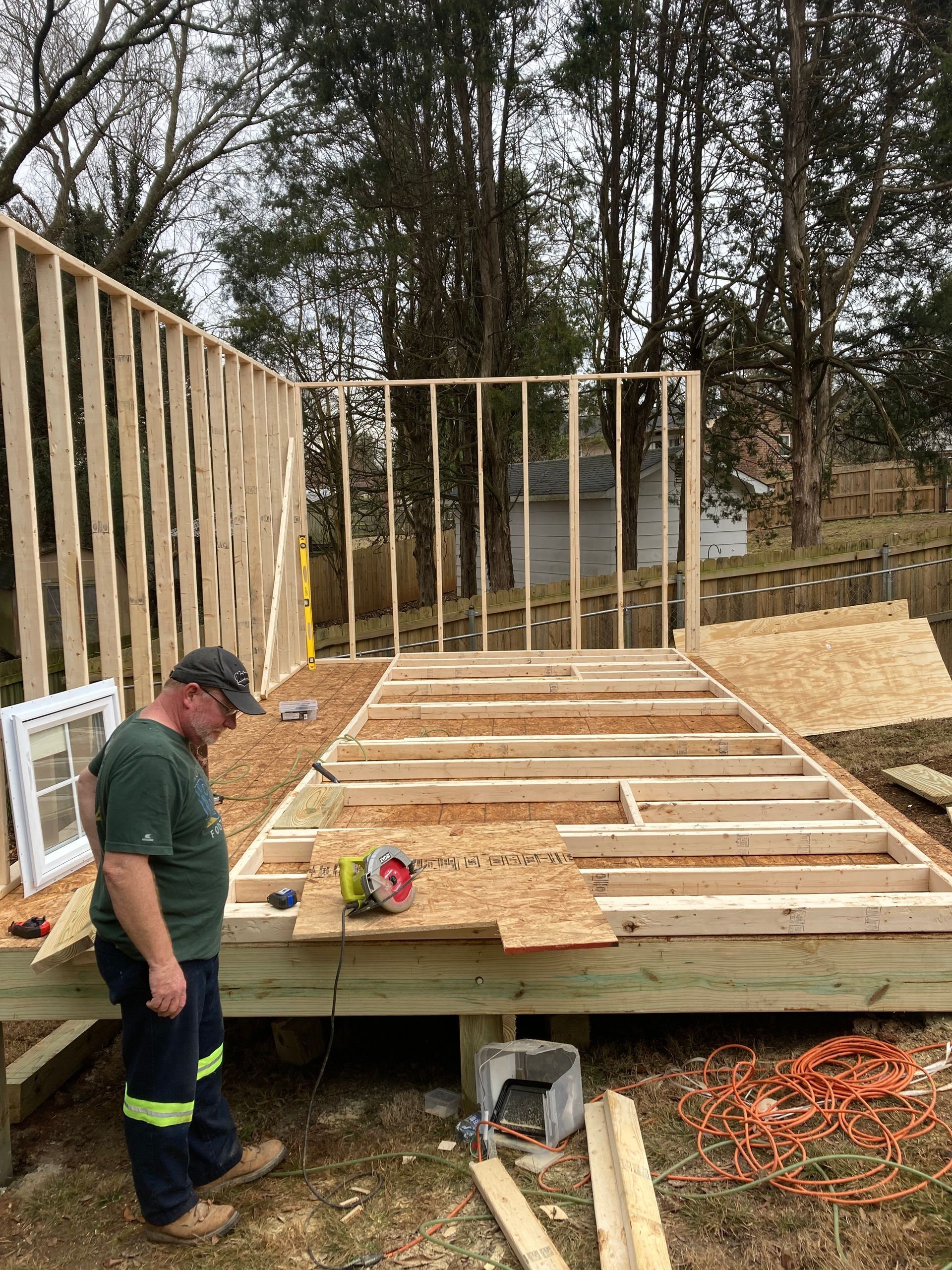 Man building a wooden structure outdoors. Saw and plywood on the deck. Framing walls are visible.