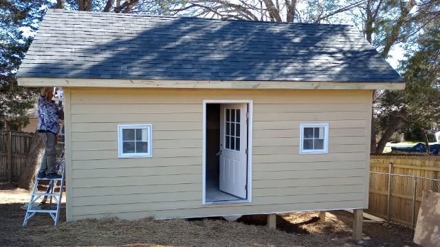 Tan shed with dark roof, white-framed door and windows. Person on ladder paints side of building.