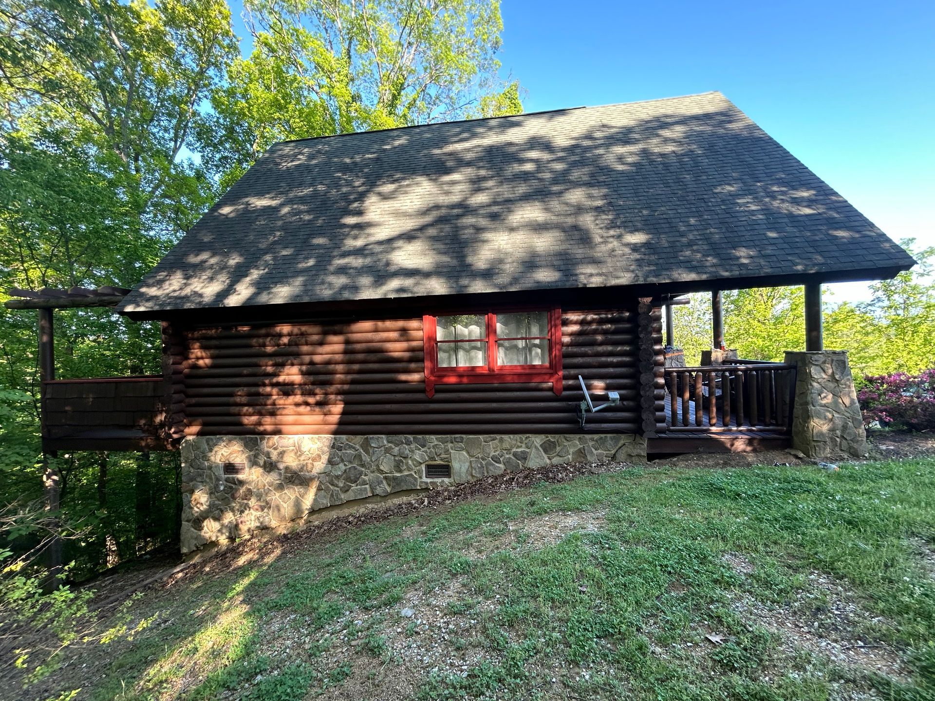 Log cabin with dark brown logs, stone foundation, red window trim, and a porch.