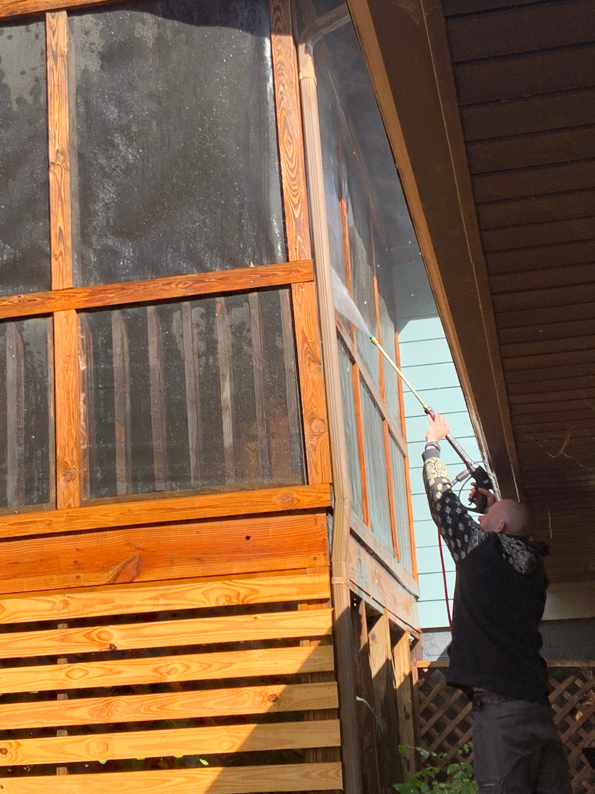 Person using a pressure washer on a wooden porch structure with windows.