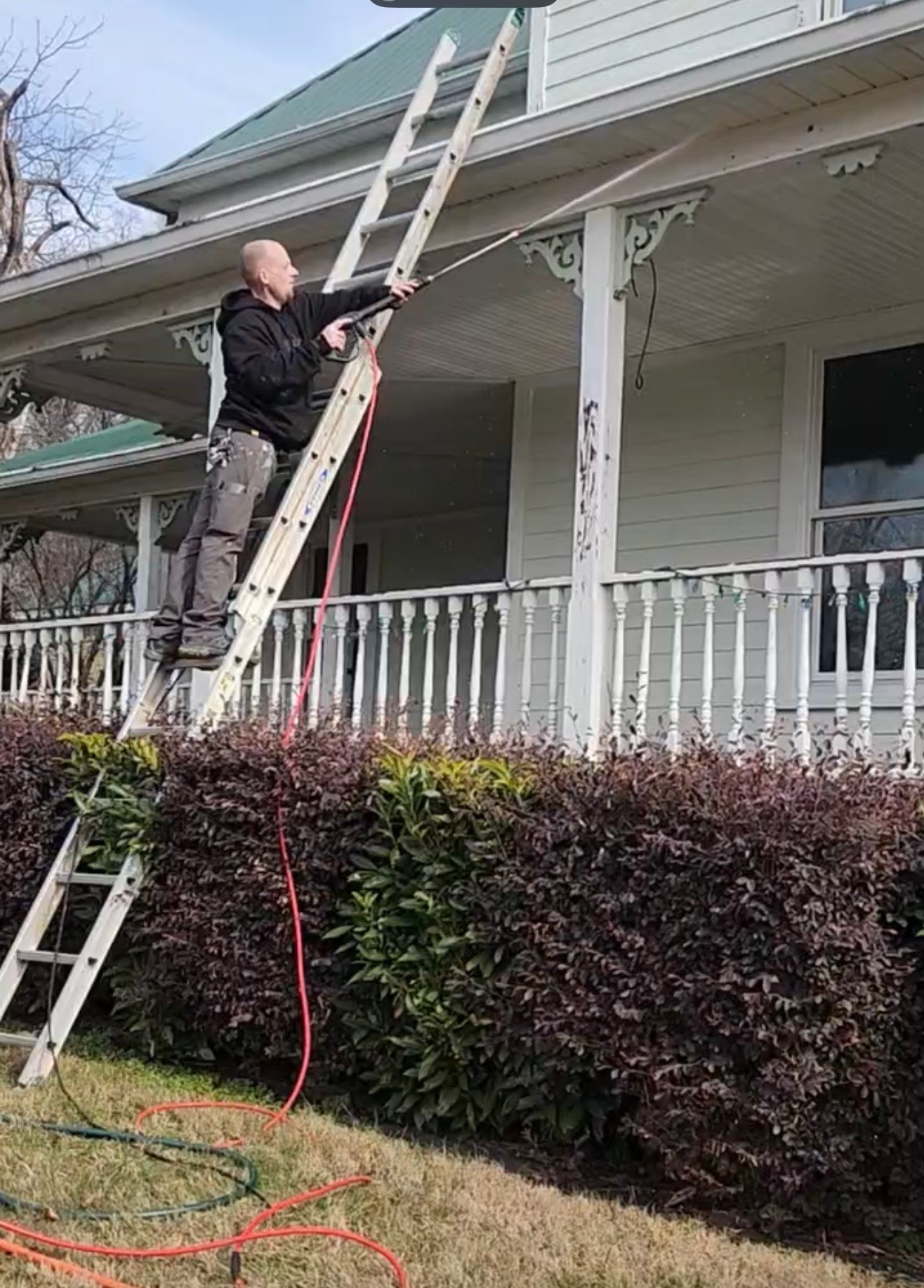 A person on a ladder cleaning a house's eaves with a pressure washer.
