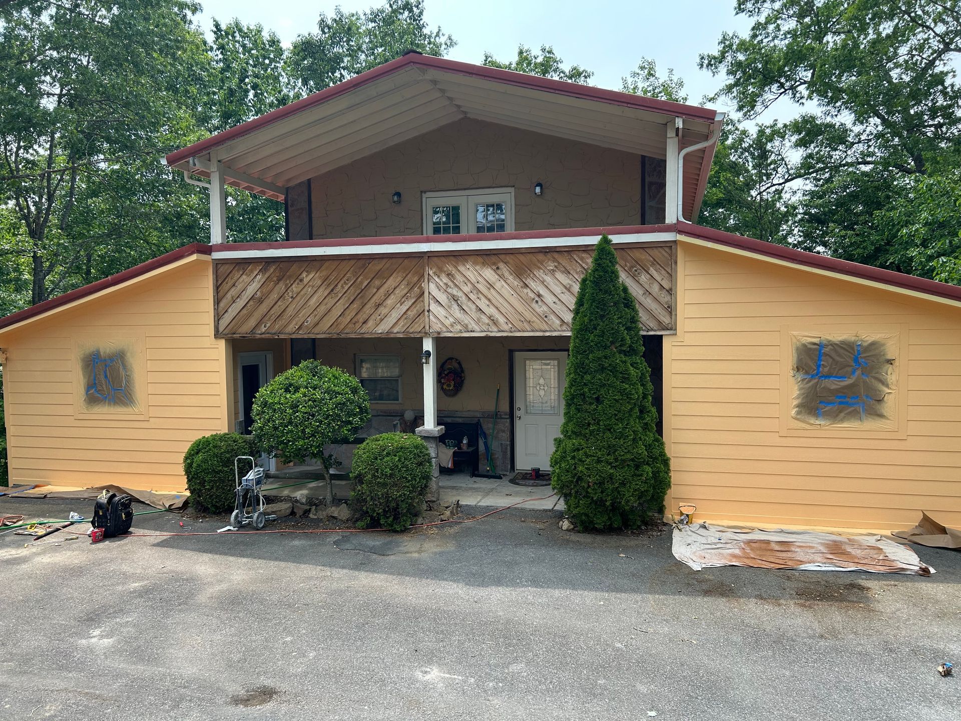Two-story tan and brown house with yellow siding and red roof, with trees and bushes in front.