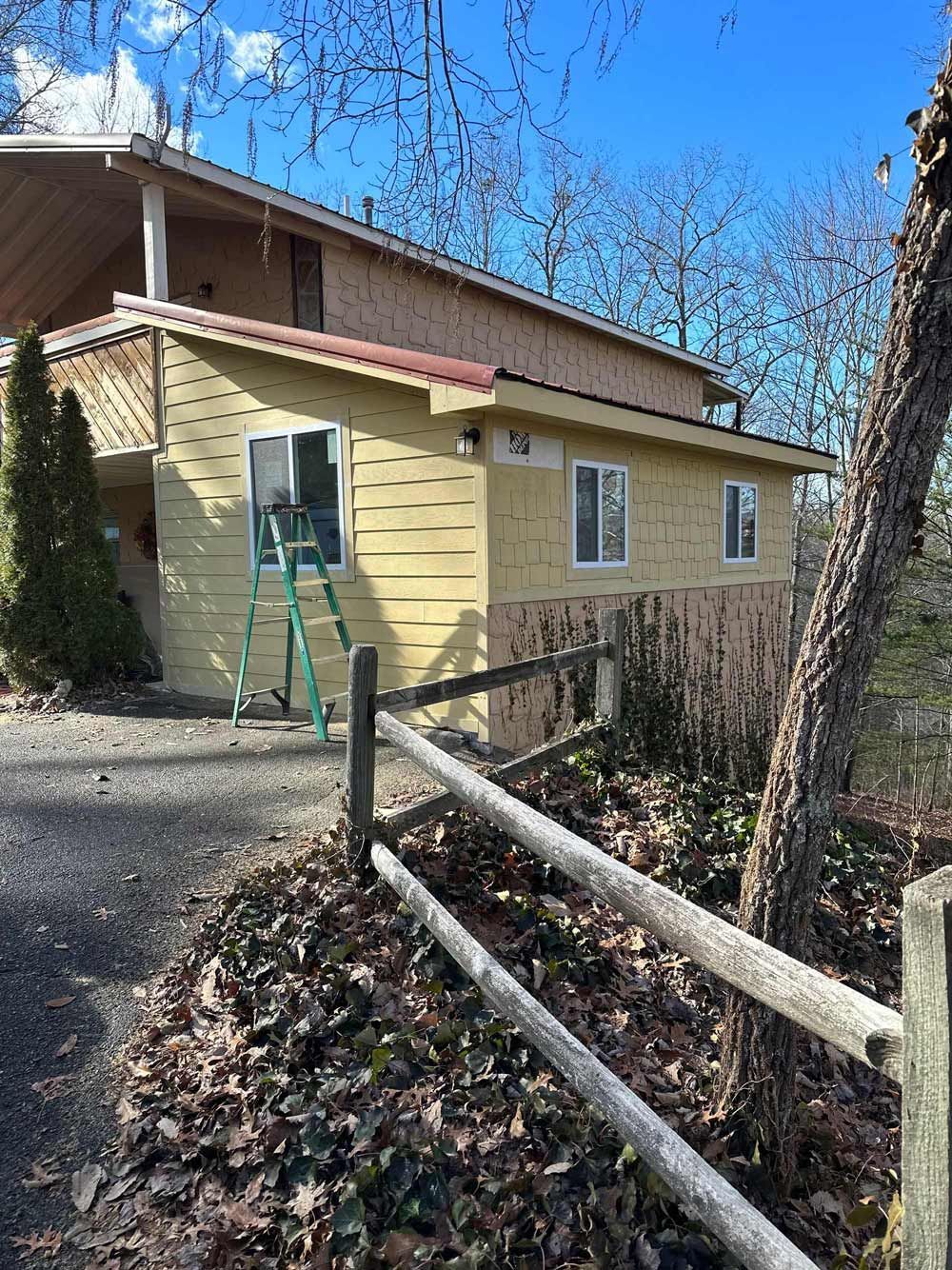 Yellow building with three windows, a ladder, and a wooden fence. Bare trees in the background.