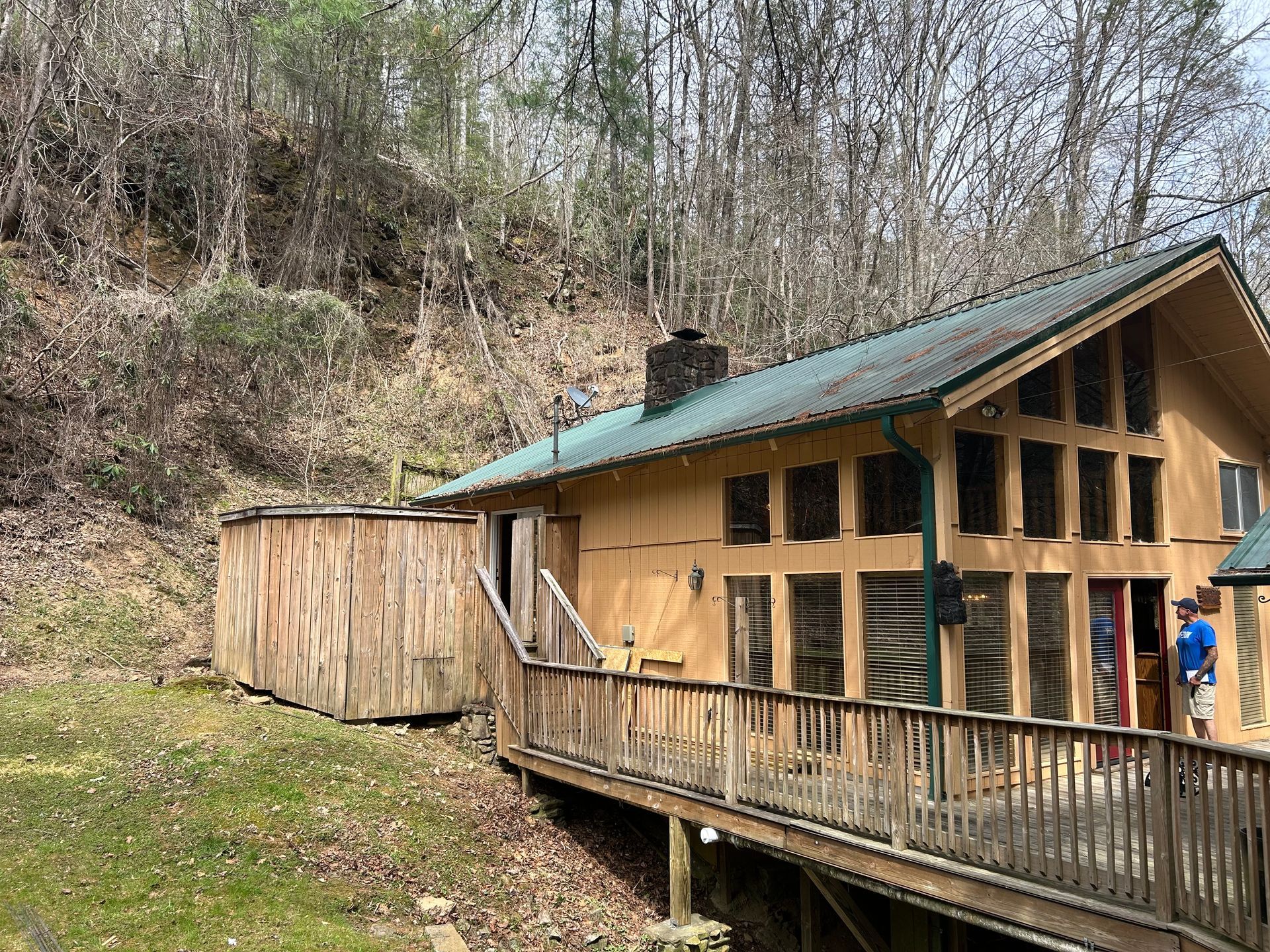 Cabin with wooden siding and green roof, deck in front. A shed sits on the side, hillside behind.