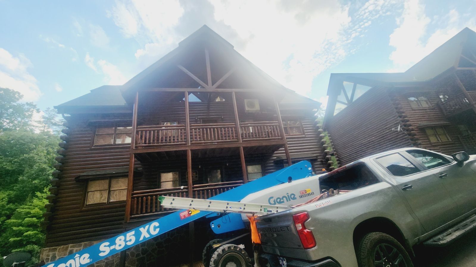 Log cabin with wooden exterior. A Genie S-85 XC lift is in use with a truck on the right. Blue sky.
