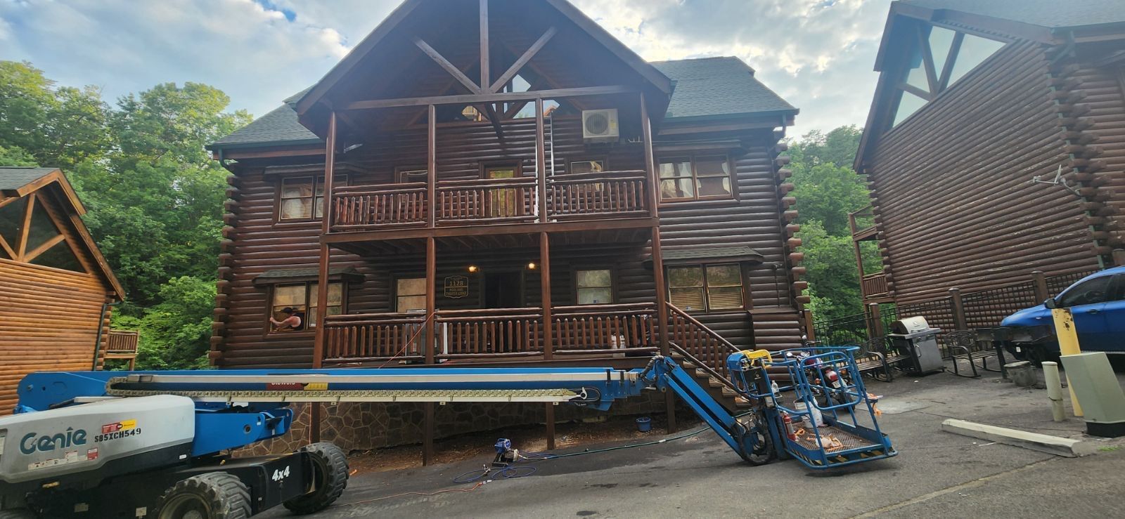 A two-story log cabin being worked on by a Genie lift. A blue truck is parked nearby.