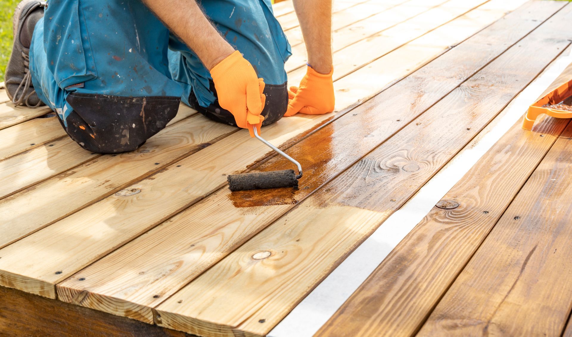 Person applying stain to a wooden deck with a roller, wearing orange gloves.