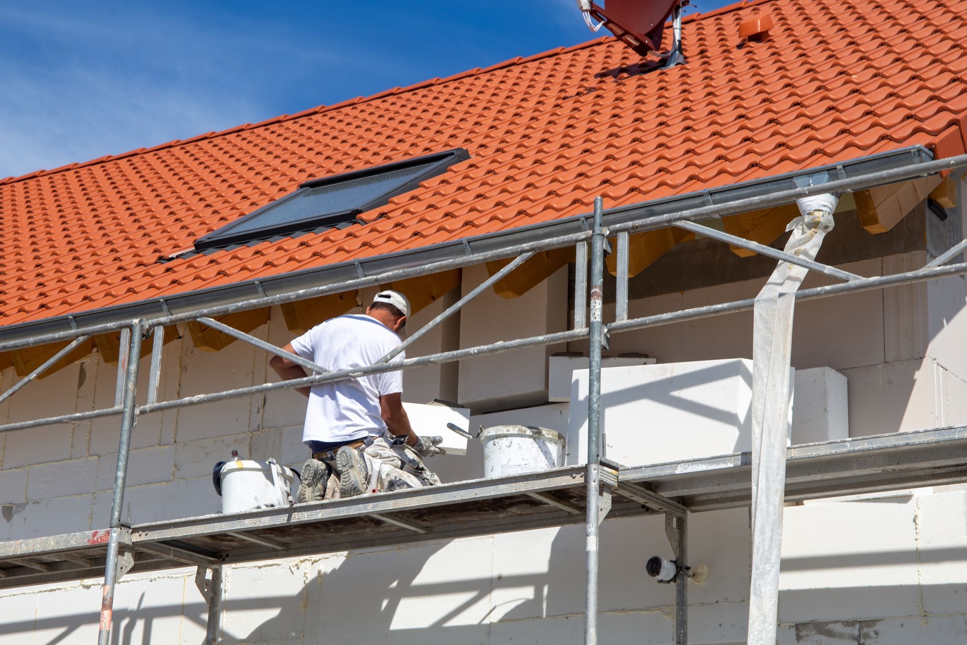 Construction worker on scaffolding, applying material to the white exterior of a building with a red tiled roof.