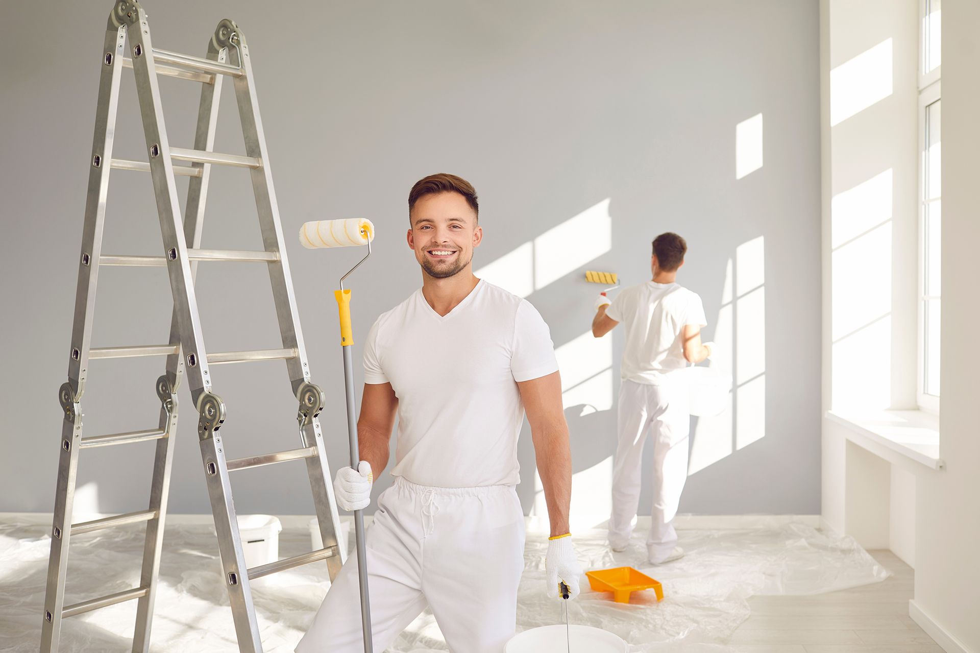 Two people painting a gray wall; one smiling, holding a paint roller; ladder nearby, sunny room.