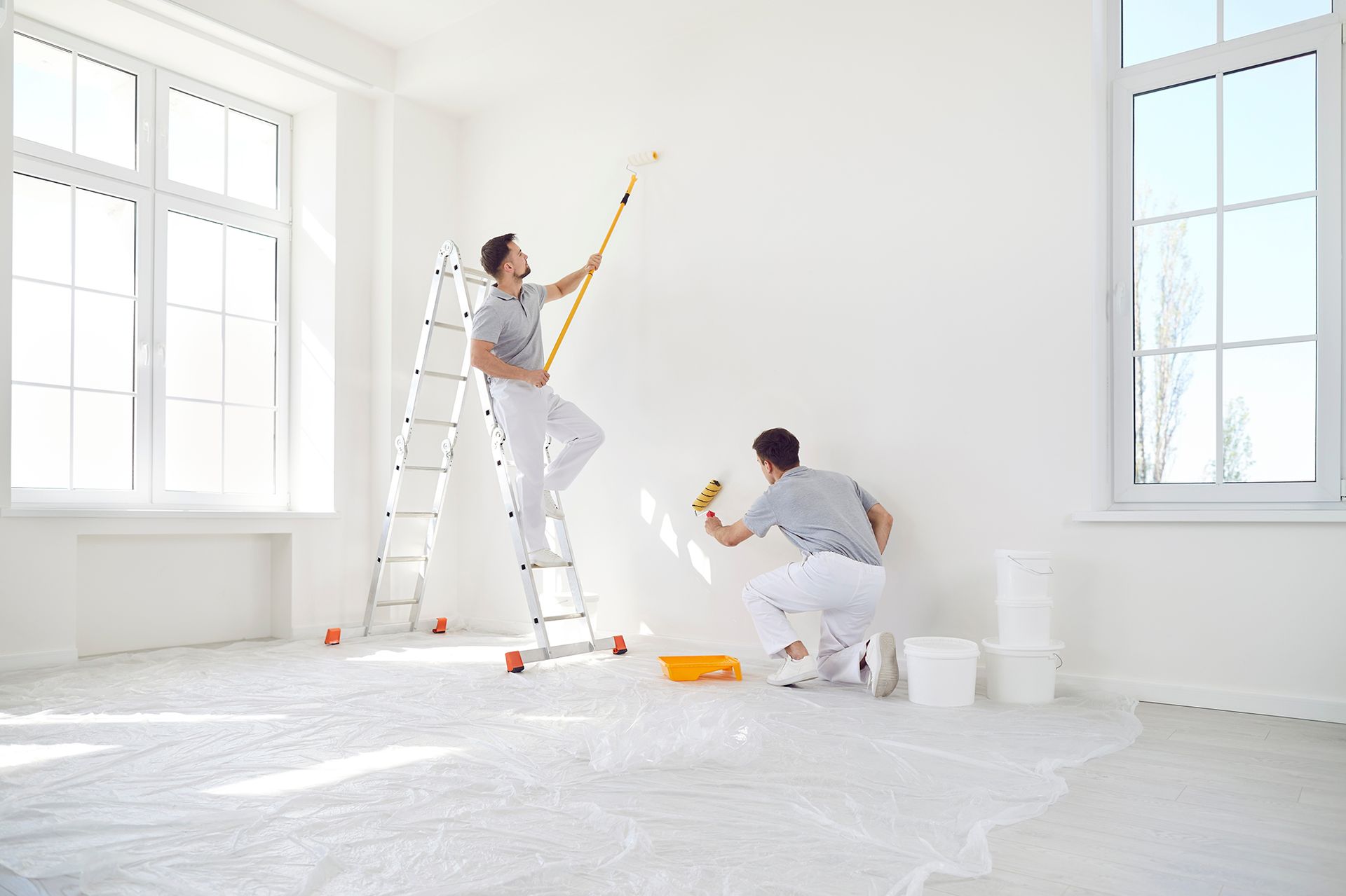 Two people painting a white wall in a room with large windows. One uses a roller, the other a brush.