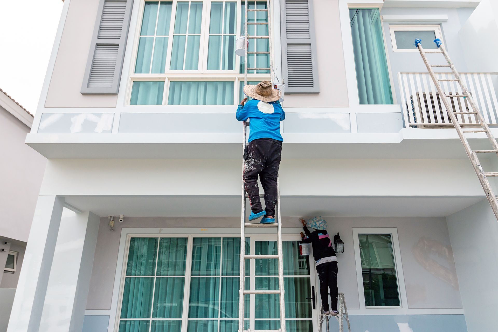 Two painters on ladders, painting exterior trim of a two-story house.