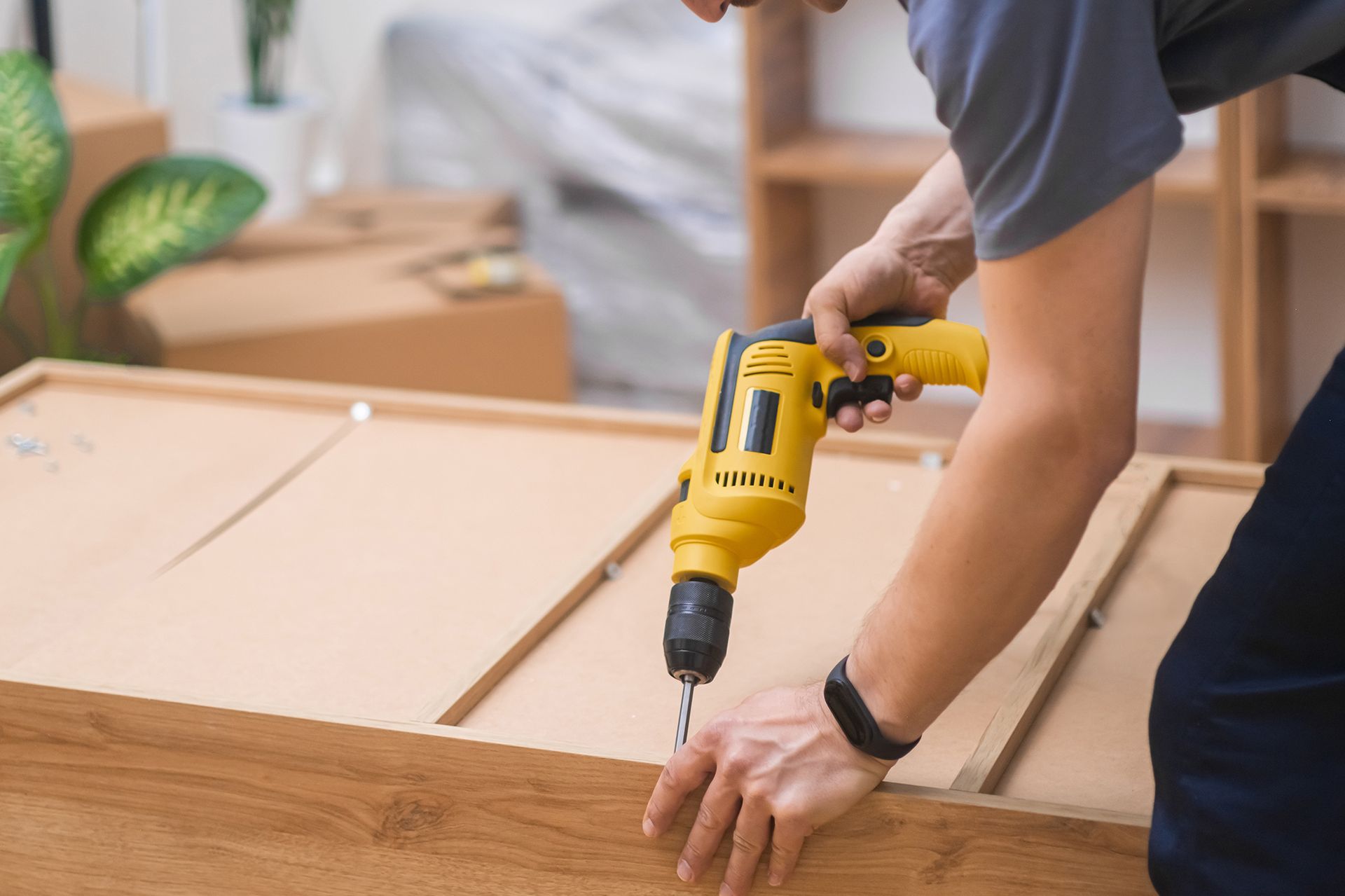 Person using a yellow drill to assemble wooden furniture in a room.