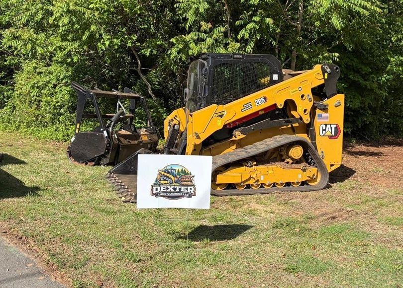 Yellow track skid steer parked on grass beside a brush cutter, with a sign in front and trees behind
