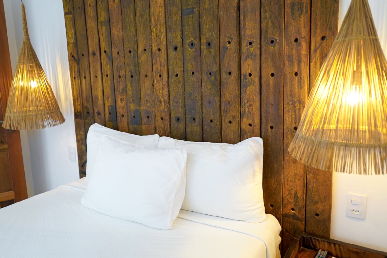 Bed with white linens, two pillows, and a rustic wooden headboard, flanked by woven pendant lights.