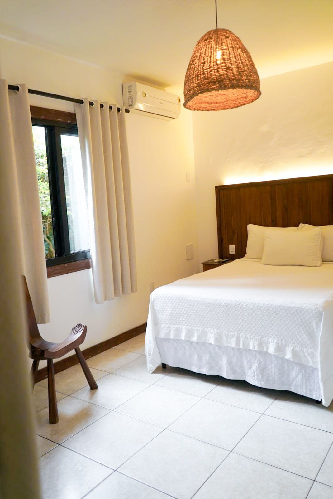 Bedroom with white bed, wooden headboard, chair, window, and woven pendant light.