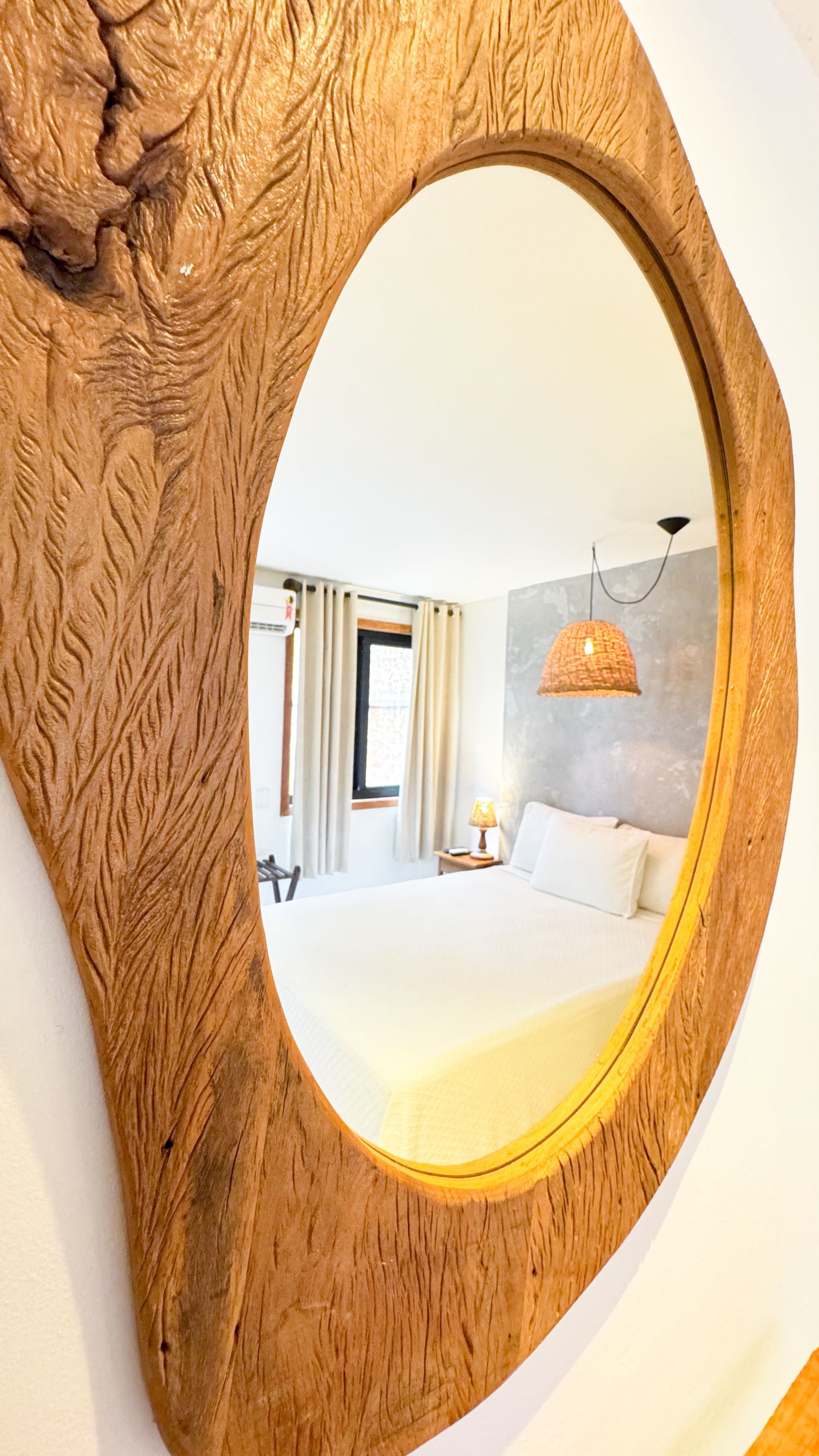 Mirror reflecting a light-filled bedroom with a bed, window, and lamp. The mirror has a wooden, organic-shaped frame.