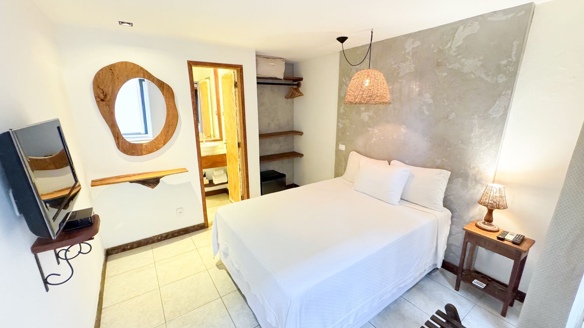 Small, modern bedroom with bed, TV, and open bathroom. White and gray walls, wooden accents, neutral colors.