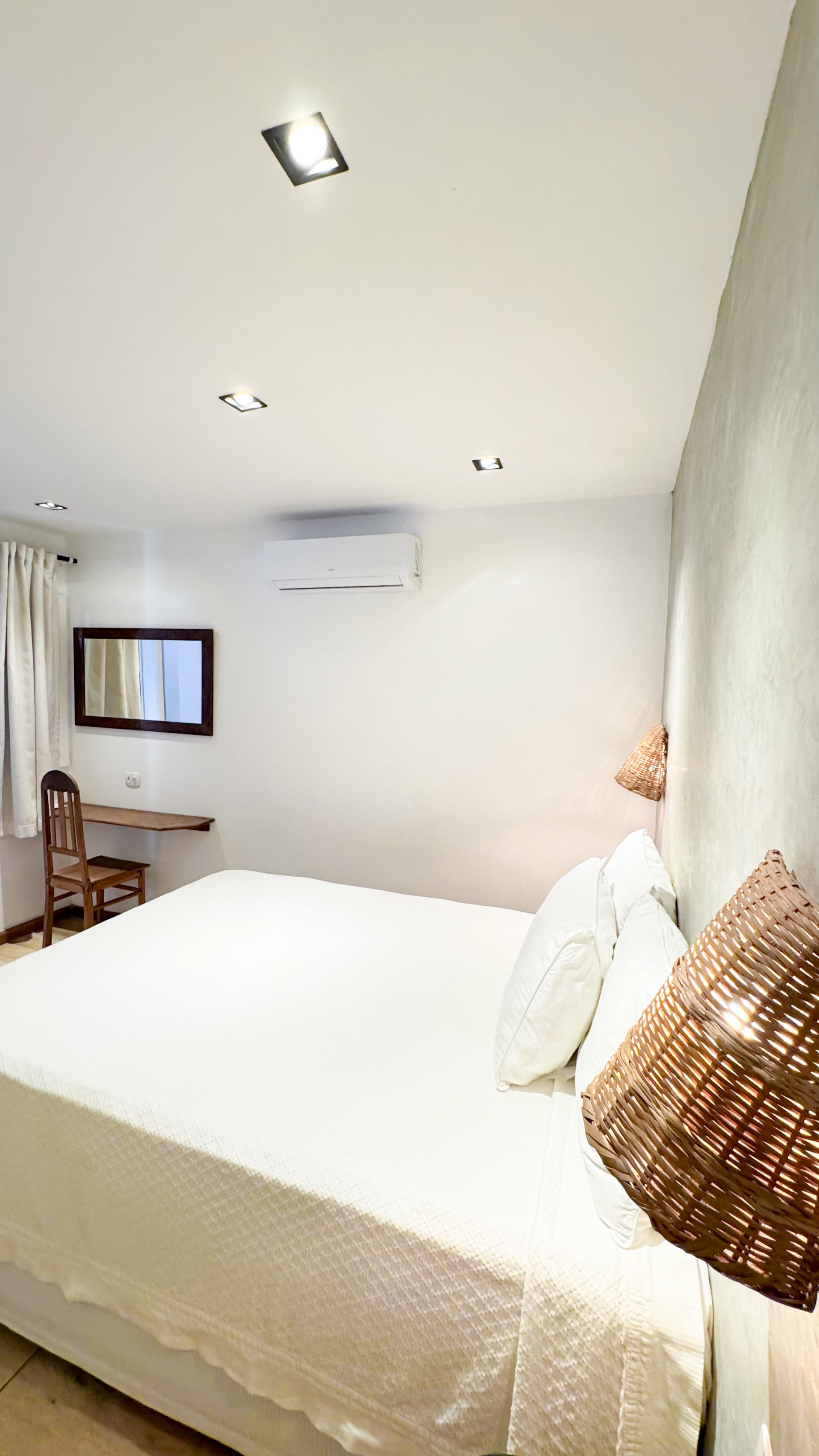 Bedroom with large bed, white linens, and decorative pillows. Desk and mirror on wall, warm lighting.