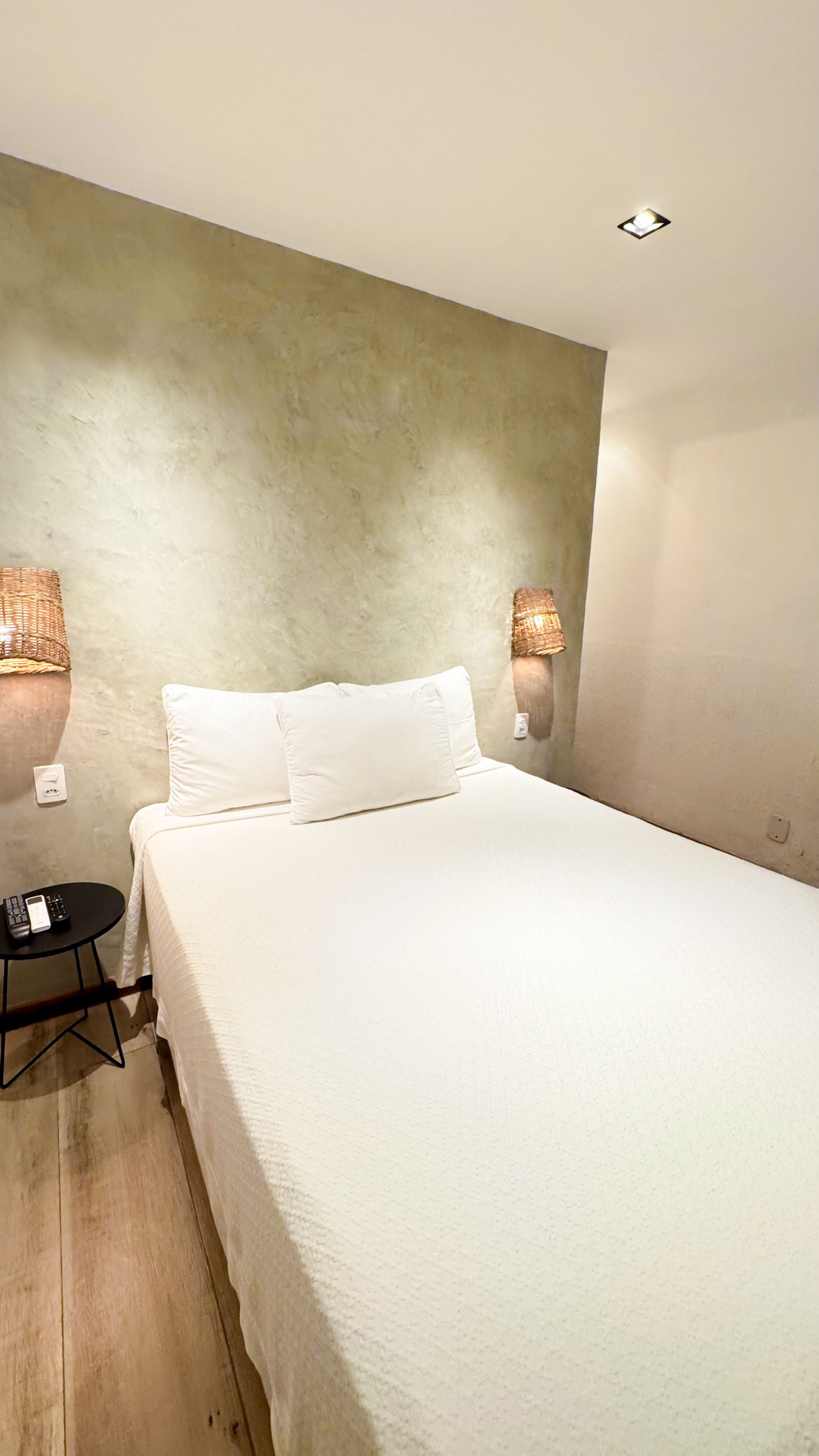A bedroom with a white bed, beige wall, and small black side table. Two wall lights illuminate the bed.