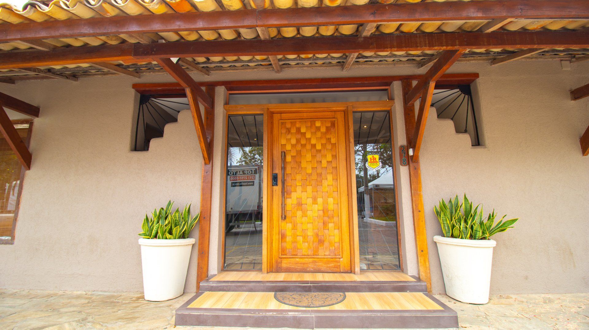 Wooden front door with glass sidelights; two white potted plants flank the entrance under a wooden awning.