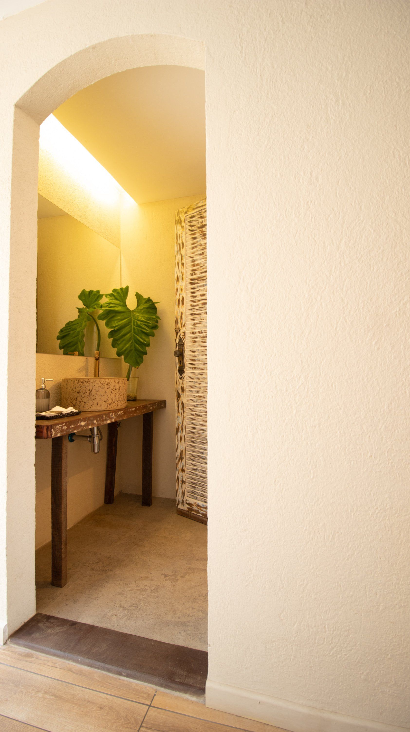 Archway leading to a bathroom; textured walls, wooden table with plant and sink, mosaic tiled wall.