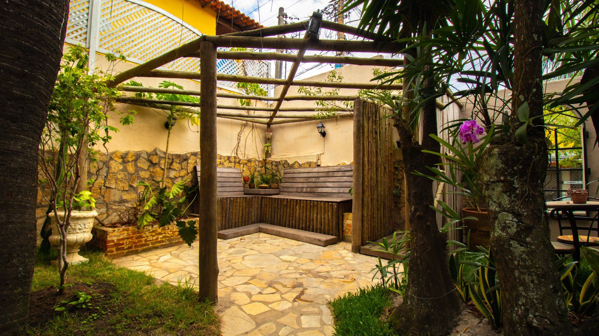 A rustic outdoor seating area with a wooden pergola, stone floor, and surrounding greenery.