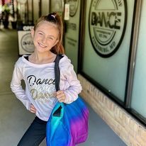 A young girl is standing in front of a dance studio holding a backpack.