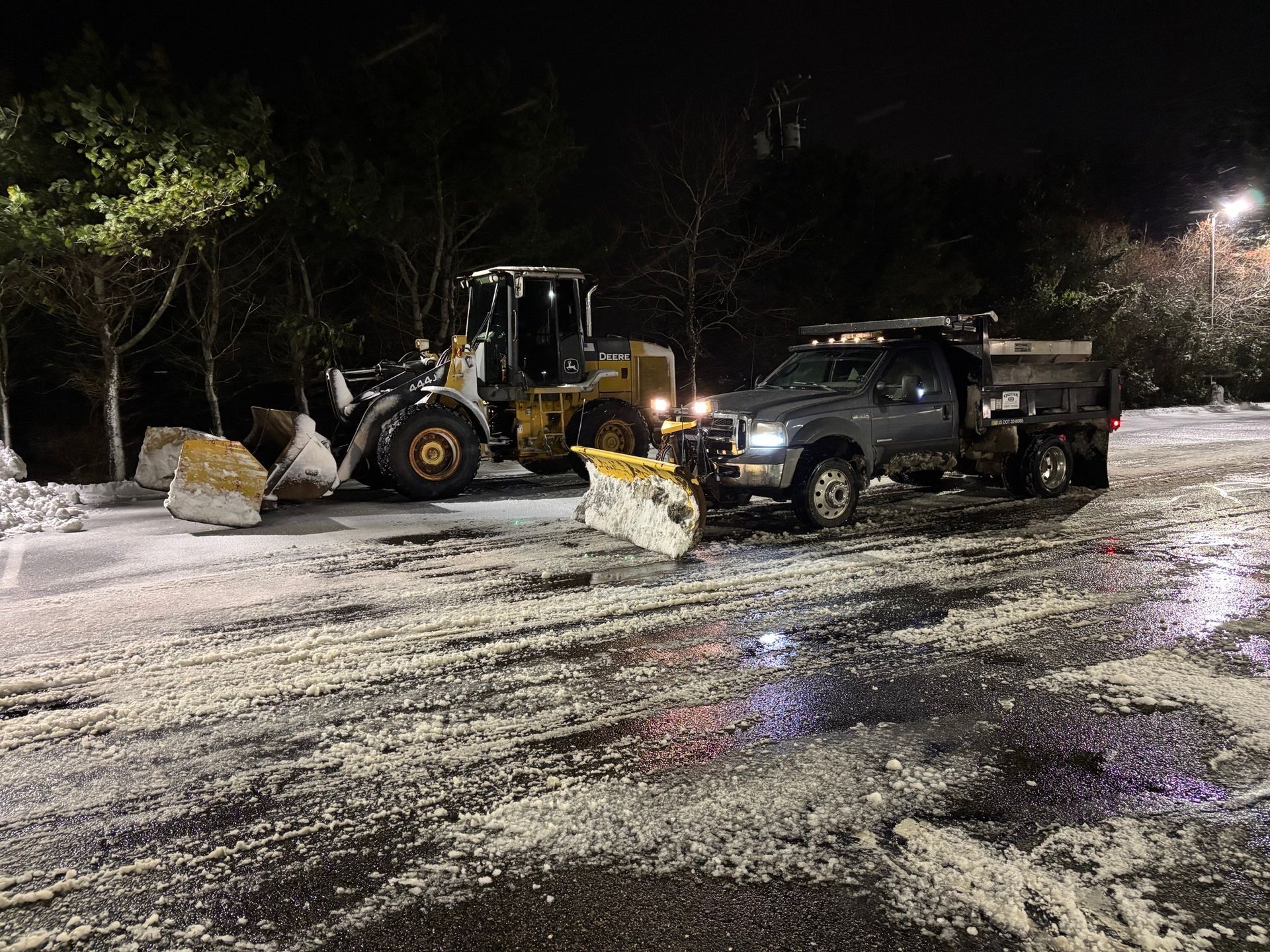 Snowplows clearing a parking lot at night. A yellow loader pushes snow; a truck with plow on the right.