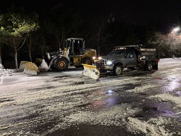 Snowplows clearing a parking lot at night. A yellow loader pushes snow; a truck with plow on the right.
