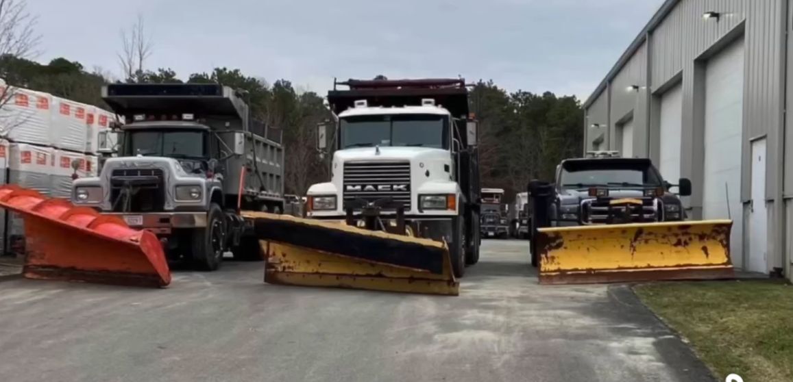 Three snowplow trucks parked on a gray surface in front of a building; orange and yellow plows.