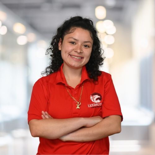 Woman with curly hair in red polo, arms crossed, smiles in a hallway.