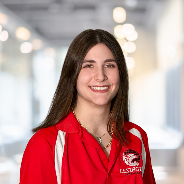 Woman with long brown hair smiles in a red Lexington polo shirt, blurred background.
