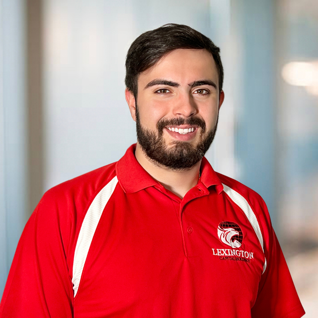 Smiling man in red polo shirt with beard, Lexington logo, blurred background.
