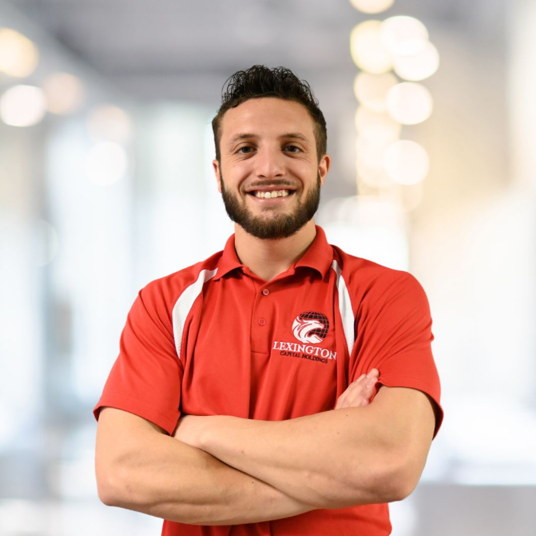 Man in red shirt with arms crossed, smiling. Blurred office background.