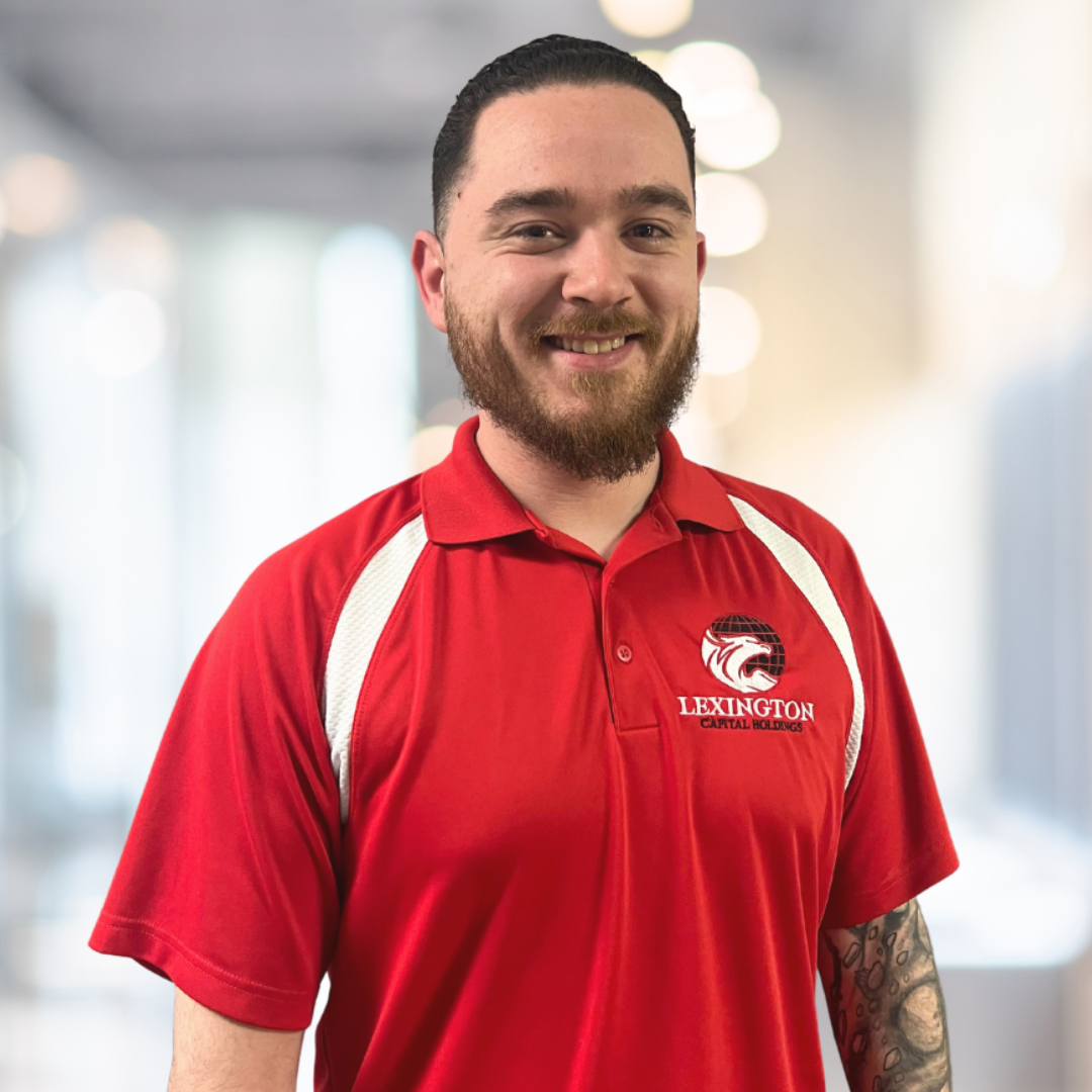 Man wearing a red Lexington polo shirt smiles at the camera; blurred office background.