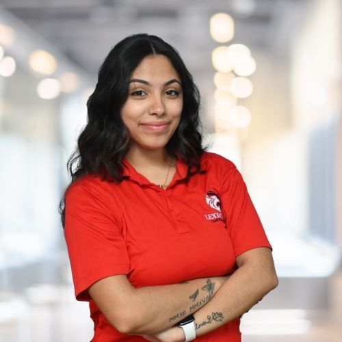 Woman with dark hair, wearing red polo shirt, arms crossed, smiling. Blurred background.
