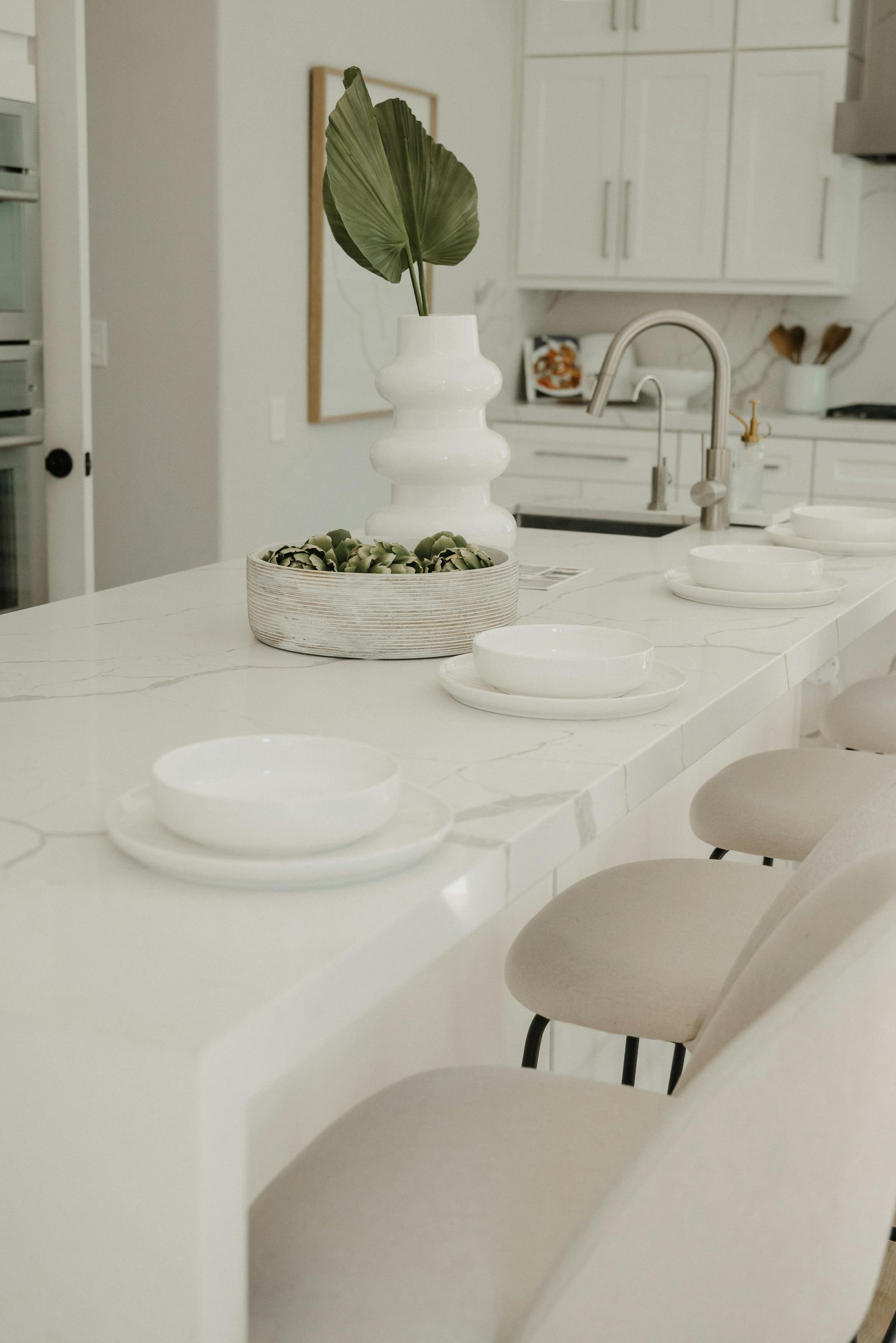 White kitchen island with chairs, serving dishes, and a vase with a large green leaf.