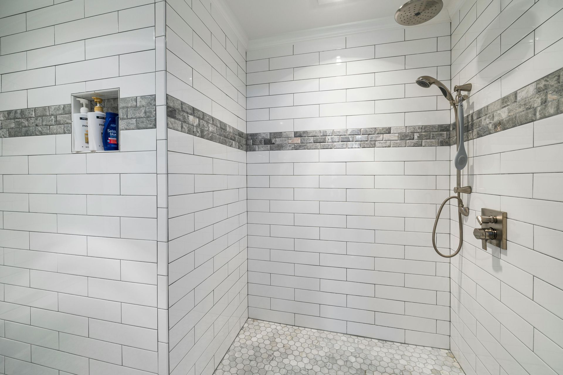 White-tiled shower with a pebble floor and grey accent tile. Features a shower head and bottles in a built-in shelf.
