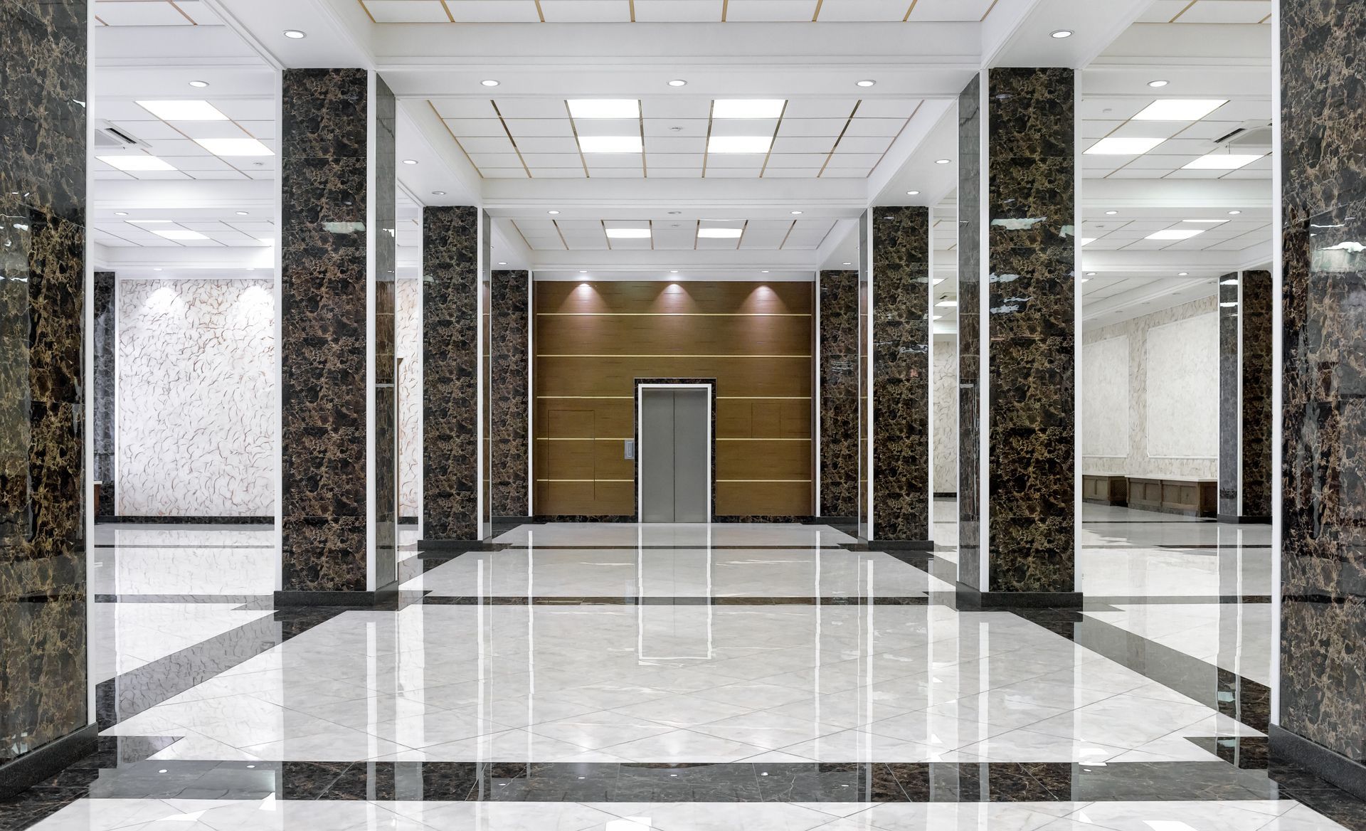 Spacious lobby with marble floors and columns, elevator at the end. Bright lighting.