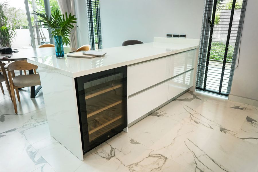 Modern white kitchen island with a built-in wine cooler, marble floors, and windows.