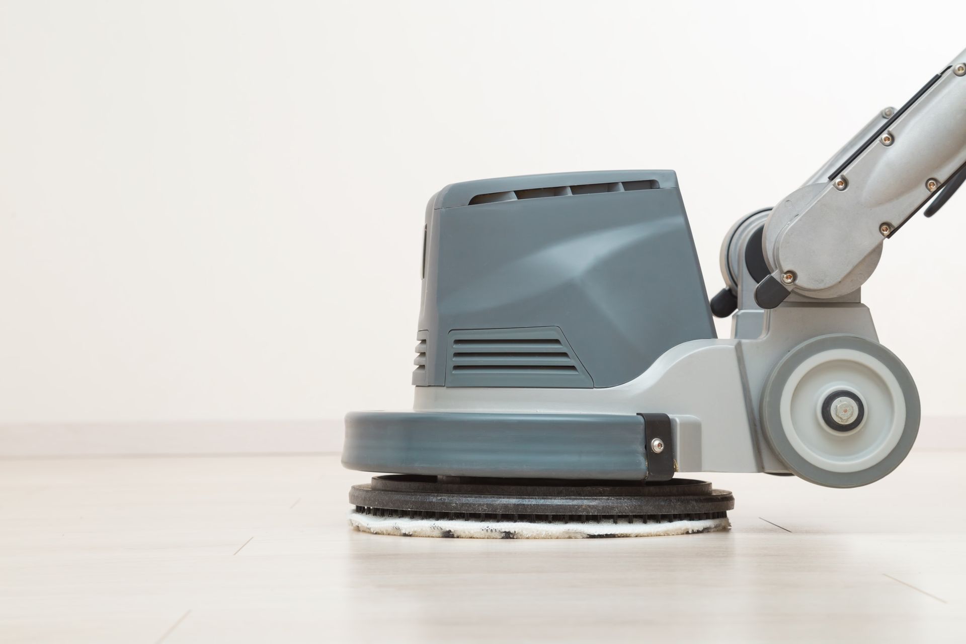 Floor cleaning machine scrubbing a light-colored wooden floor in a white room.