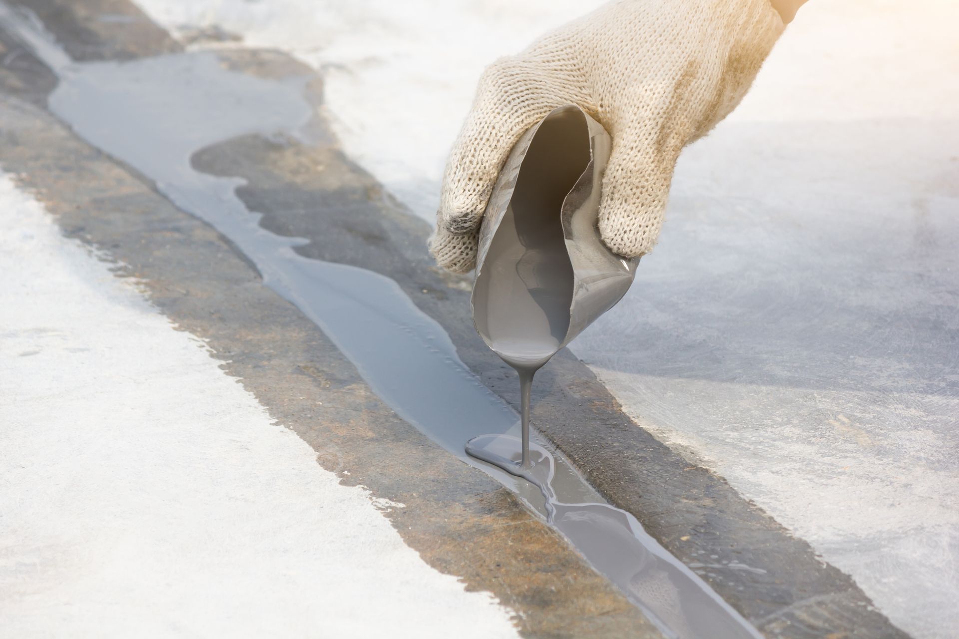 Gloved hand pouring gray sealant into a concrete joint. Sunlight shines on the surface.