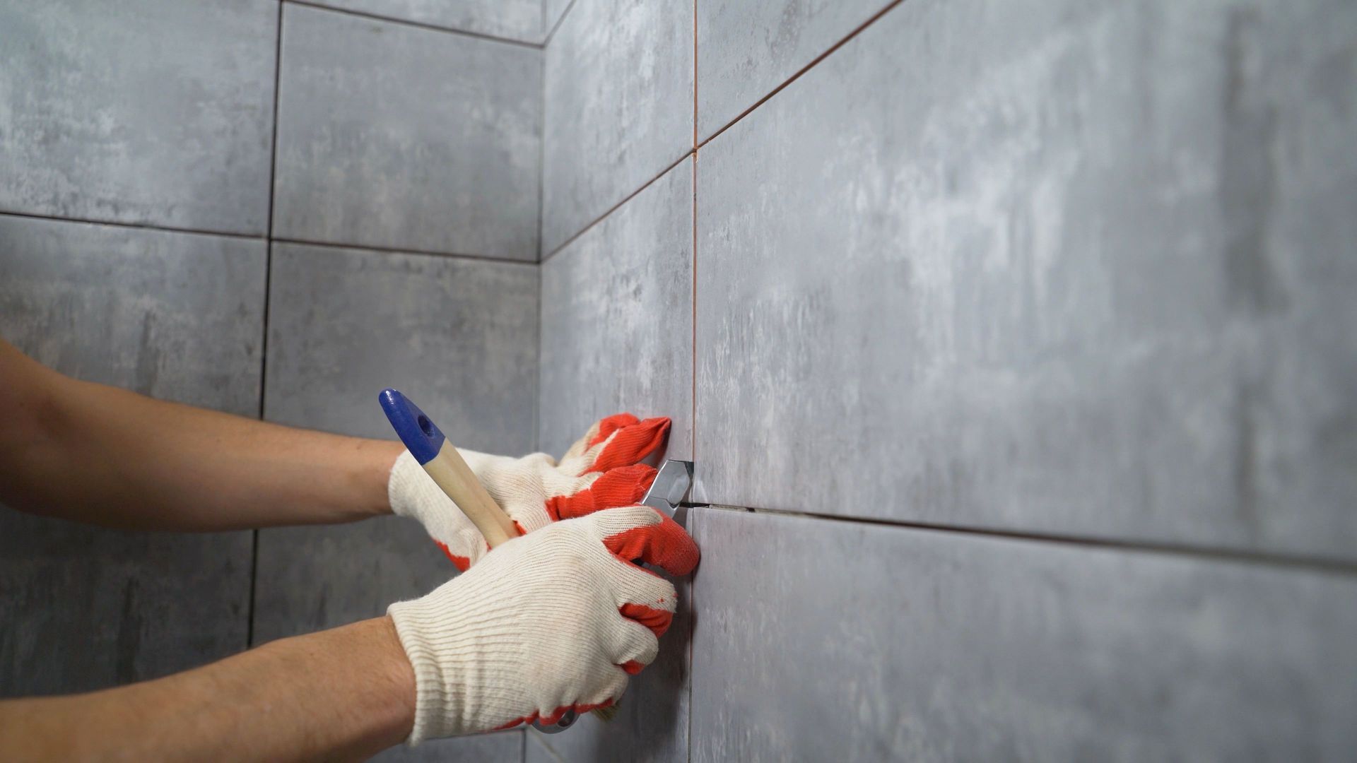 Person wearing gloves using a tool to work on gray tile in a bathroom.