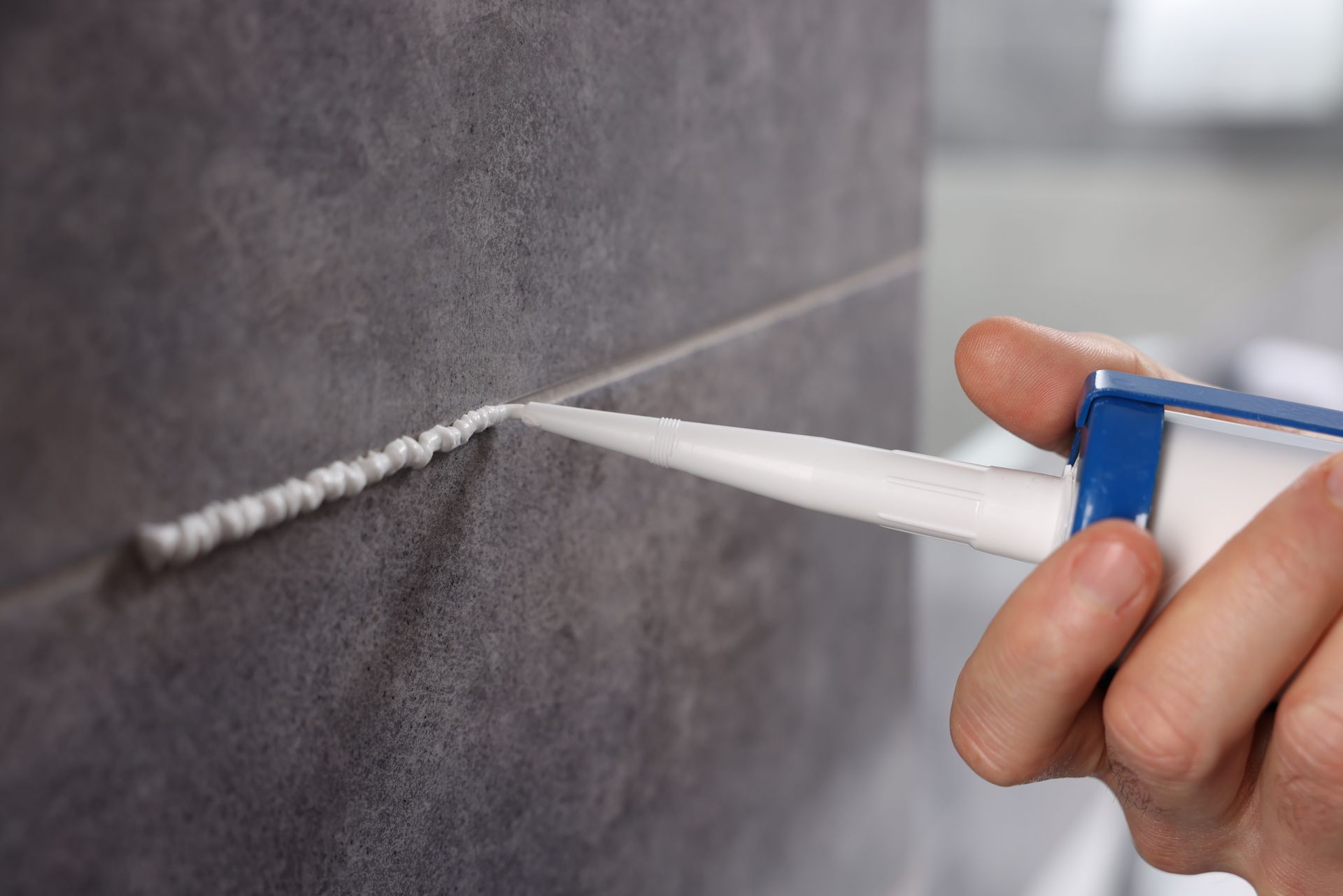 Hand applying white caulk between dark gray tiles.