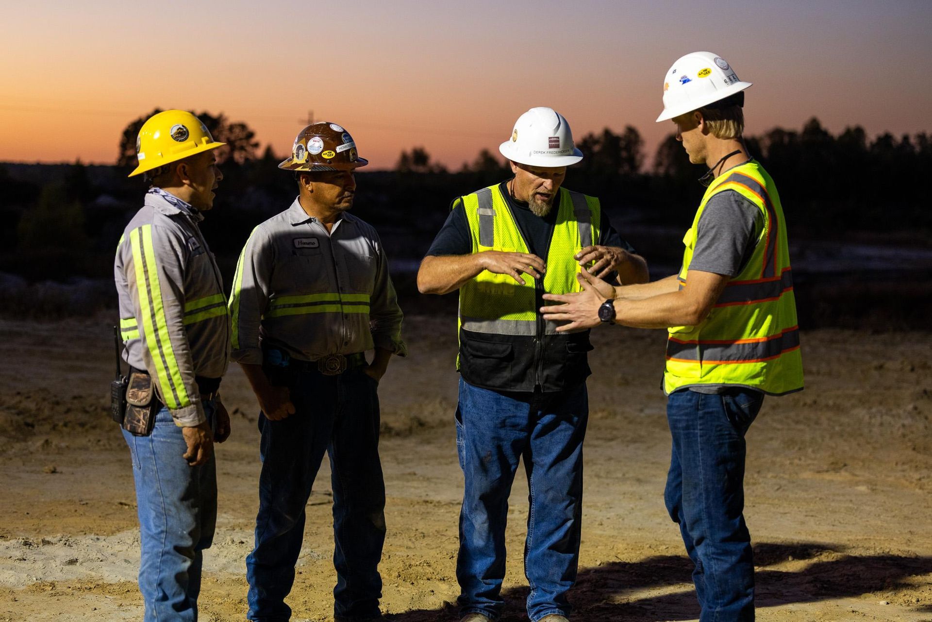 Two construction workers in hard hats and vests consult near a large sand pile.