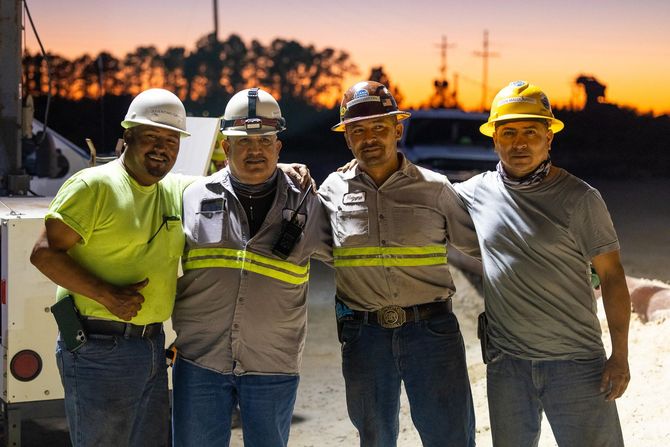 Construction site: Operator in excavator cab, worker in safety vest outside, dump truck nearby.