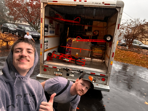 Two men are posing for a picture in front of a moving truck.