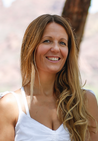 Woman smiles, long blonde hair, white top, outdoors with blurred background.
