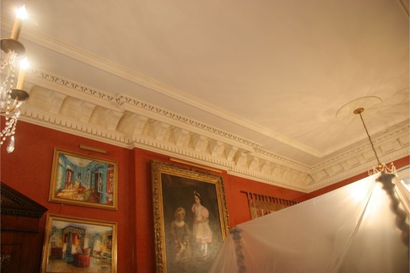 Red walls, ornate white ceiling, framed paintings, and a draped spotlight in a gallery-like room